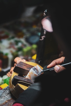 A person engraves intricate designs on a small object held in a vise. The scene is dimly lit, highlighting the tools and the skillful hand at work, suggesting a craft or artisanal activity.