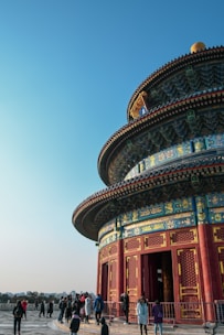 A vibrant, ornately decorated round temple structure with intricate details and rich colors stands prominently against a clear blue sky. A group of people, likely tourists, gather around the temple, adding a sense of activity and scale to the scene.