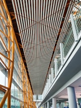 A modern architectural interior featuring a high ceiling with parallel wooden slats running lengthwise. The structure includes golden metal beams on the left supporting large glass windows, providing natural light. The right side showcases a glass railing on an upper level with white columns supporting the structure below.