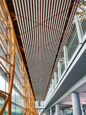 A modern architectural interior featuring a high ceiling with parallel wooden slats running lengthwise. The structure includes golden metal beams on the left supporting large glass windows, providing natural light. The right side showcases a glass railing on an upper level with white columns supporting the structure below.