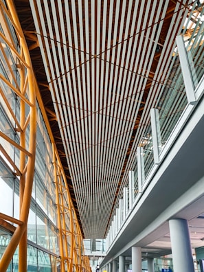 A modern architectural interior featuring a high ceiling with parallel wooden slats running lengthwise. The structure includes golden metal beams on the left supporting large glass windows, providing natural light. The right side showcases a glass railing on an upper level with white columns supporting the structure below.