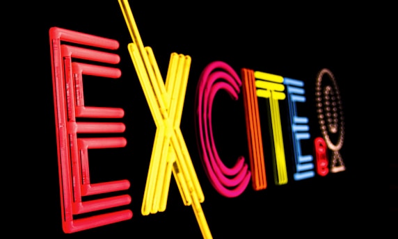 Bright neon letters spell out the word 'EXCITE' against a black background, using a variety of colors including red, yellow, pink, and blue.