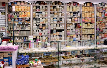 A vibrant and colorful shop filled with shelves stacked high with rolls of fabric and lace. The shelves display a variety of intricate patterns and colors, showcasing diverse textile options. Below the shelves is a glass counter containing trays with small various items. The labels above the shelves and the bustling layout suggest a fabric or craft store specializing in embroidery and lace.