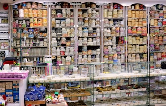 A vibrant and colorful shop filled with shelves stacked high with rolls of fabric and lace. The shelves display a variety of intricate patterns and colors, showcasing diverse textile options. Below the shelves is a glass counter containing trays with small various items. The labels above the shelves and the bustling layout suggest a fabric or craft store specializing in embroidery and lace.