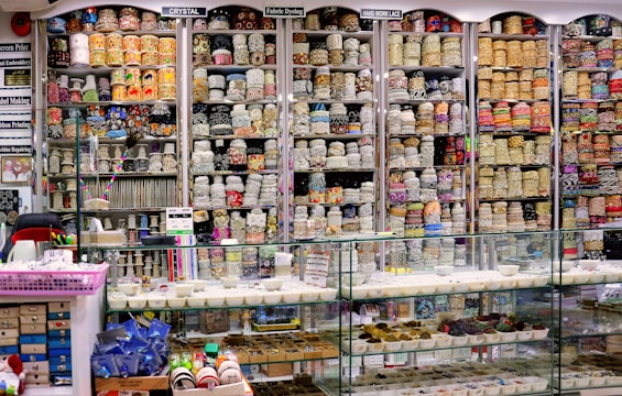 A vibrant and colorful shop filled with shelves stacked high with rolls of fabric and lace. The shelves display a variety of intricate patterns and colors, showcasing diverse textile options. Below the shelves is a glass counter containing trays with small various items. The labels above the shelves and the bustling layout suggest a fabric or craft store specializing in embroidery and lace.
