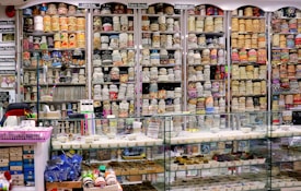 A vibrant and colorful shop filled with shelves stacked high with rolls of fabric and lace. The shelves display a variety of intricate patterns and colors, showcasing diverse textile options. Below the shelves is a glass counter containing trays with small various items. The labels above the shelves and the bustling layout suggest a fabric or craft store specializing in embroidery and lace.