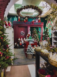 A festive Christmas shop decorated with a variety of holiday ornaments and greenery. The entrance is adorned with a garland and a large wreath hangs on a door. Several Christmas trees are spread throughout, each elaborately decorated with baubles and lights. The store is filled with Christmas decorations and figurines, with a focus on vibrant reds, greens, and golds.