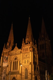 A Gothic-style cathedral with two prominent spires illuminated at night, showcasing intricate architectural details and a banner promoting refugee support hanging on the facade.