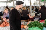 A man wearing a black coat and cap is holding a squash while standing in an outdoor market. Various vegetables, including carrots and leafy greens, are displayed on the table in front of him. Other market-goers and vendors are visible in the background, with the scene set under a covered structure.