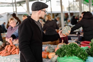 A man wearing a black coat and cap is holding a squash while standing in an outdoor market. Various vegetables, including carrots and leafy greens, are displayed on the table in front of him. Other market-goers and vendors are visible in the background, with the scene set under a covered structure.