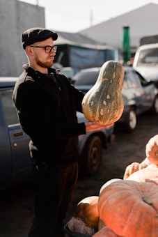 A person wearing glasses and a black cap inspects a large, uniquely shaped squash. The background includes vehicles and a market scene with sunlight illuminating the area.