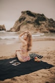 A woman in a floral swimsuit sits on a black beach blanket with a rocky ocean backdrop. Nearby are sunglasses, a tablet, and a container of strawberries on the blanket. Her hand is in her hair, and the setting evokes a relaxed beach atmosphere.