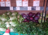 A selection of fresh vegetables is displayed, including leafy green lettuce, purple cabbages, and various leafy greens arranged neatly in baskets. Above the vegetables are price labels in a store setting.