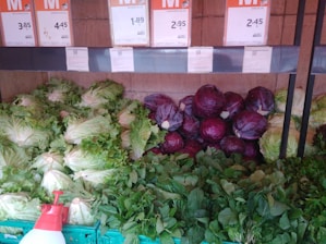A selection of fresh vegetables is displayed, including leafy green lettuce, purple cabbages, and various leafy greens arranged neatly in baskets. Above the vegetables are price labels in a store setting.