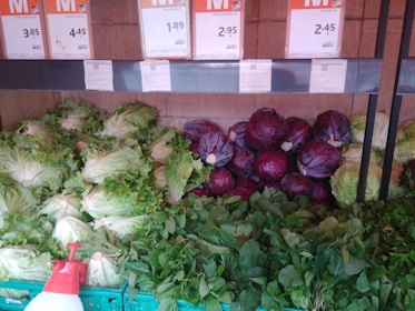 A selection of fresh vegetables is displayed, including leafy green lettuce, purple cabbages, and various leafy greens arranged neatly in baskets. Above the vegetables are price labels in a store setting.