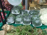 Plastic-wrapped broccoli is neatly stacked in trays, surrounded by various fresh vegetables including cauliflower and a type of root vegetable with leafy green stalks. The background features a poster with the text 'Iyi Tarim Uygulamalari' and an image of fruits.
