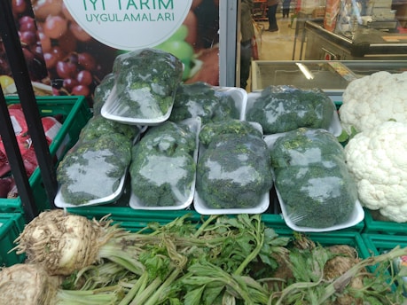 Plastic-wrapped broccoli is neatly stacked in trays, surrounded by various fresh vegetables including cauliflower and a type of root vegetable with leafy green stalks. The background features a poster with the text 'Iyi Tarim Uygulamalari' and an image of fruits.