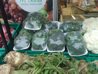 Plastic-wrapped broccoli is neatly stacked in trays, surrounded by various fresh vegetables including cauliflower and a type of root vegetable with leafy green stalks. The background features a poster with the text 'Iyi Tarim Uygulamalari' and an image of fruits.