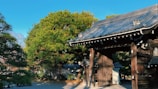 A traditional wooden gate with intricately detailed dark wood and a tiled roof stands prominently in a serene setting. Behind it, lush green trees fill the background under a clear blue sky, creating a harmonious blend of architecture and nature.