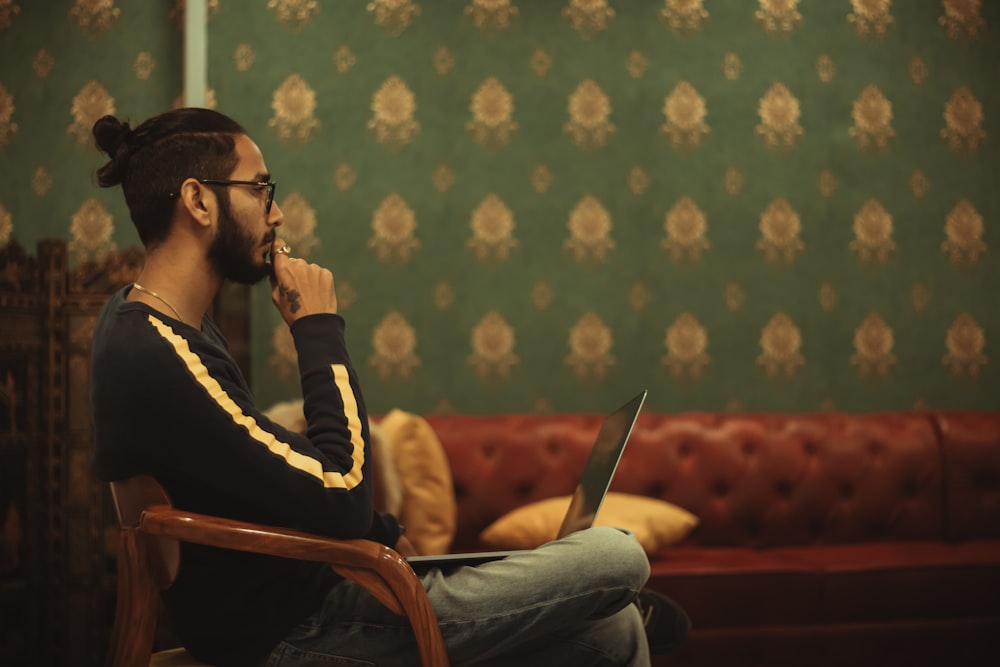 man sitting on chair while using laptop on his lap near sofa inside room