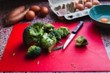 A variety of kitchen items are displayed, including a bunch of broccoli on a red cutting board with a knife beside it. In the background, there are several brown eggs with one cracked open in a blue bowl. Sweet potatoes are partially visible alongside a cardboard egg carton.