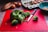 A variety of kitchen items are displayed, including a bunch of broccoli on a red cutting board with a knife beside it. In the background, there are several brown eggs with one cracked open in a blue bowl. Sweet potatoes are partially visible alongside a cardboard egg carton.