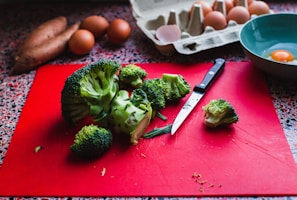 A variety of kitchen items are displayed, including a bunch of broccoli on a red cutting board with a knife beside it. In the background, there are several brown eggs with one cracked open in a blue bowl. Sweet potatoes are partially visible alongside a cardboard egg carton.