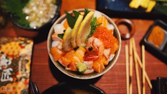 A bowl of fresh seafood and sushi toppings is garnished with lemon slices, including salmon, shrimp, squid, and salmon roe. Wasabi and green leaves accompany the dish, which is artistically presented. Surrounding the bowl are chopsticks and other dishes on a wooden table.