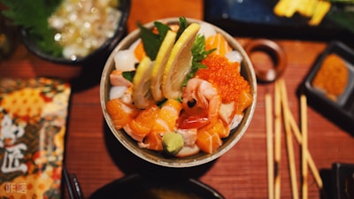A bowl of fresh seafood and sushi toppings is garnished with lemon slices, including salmon, shrimp, squid, and salmon roe. Wasabi and green leaves accompany the dish, which is artistically presented. Surrounding the bowl are chopsticks and other dishes on a wooden table.