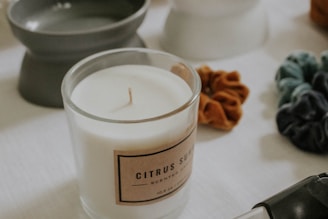 A glass jar candle labeled 'Citrus Sume' is placed on a white surface. Next to it, several fabric scrunchies and a soft-focus gray container are visible, suggesting a cozy or self-care setting.