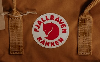 A brown fabric item with two woven straps and a circular logo in the center. The logo features a red stylized fox and text, set against a white background.