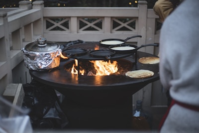 A round outdoor grill with a central open flame is being used. Multiple frying pans, each holding batter, are positioned on the grill, likely for making crepes or pancakes. A pot wrapped in aluminum foil sits nearby. The scene is outdoors, with a wooden fence and soft, ambient lighting visible in the background.