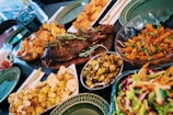 A lavish spread of various dishes including a whole roasted fish garnished with herbs, roasted potatoes, a colorful salad, glazed Brussels sprouts, and a bowl of golden-brown roasted vegetables. The dishes are served on a wooden table with green plates and utensils.