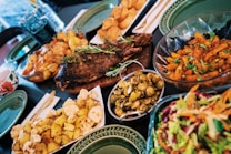 A lavish spread of various dishes including a whole roasted fish garnished with herbs, roasted potatoes, a colorful salad, glazed Brussels sprouts, and a bowl of golden-brown roasted vegetables. The dishes are served on a wooden table with green plates and utensils.