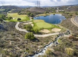 A scenic landscape featuring a golf course integrated into a hilly terrain with lush green fairways and sand traps. There is a large, tranquil lake reflecting the sky, surrounded by various trees. A winding path runs through the landscape, and power lines stretch across the hills in the background.