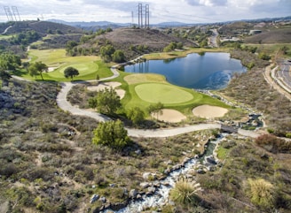 A scenic landscape featuring a golf course integrated into a hilly terrain with lush green fairways and sand traps. There is a large, tranquil lake reflecting the sky, surrounded by various trees. A winding path runs through the landscape, and power lines stretch across the hills in the background.