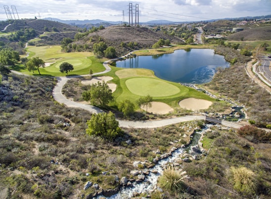 A scenic landscape featuring a golf course integrated into a hilly terrain with lush green fairways and sand traps. There is a large, tranquil lake reflecting the sky, surrounded by various trees. A winding path runs through the landscape, and power lines stretch across the hills in the background.
