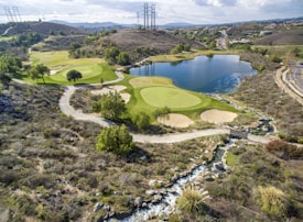 A scenic landscape featuring a golf course integrated into a hilly terrain with lush green fairways and sand traps. There is a large, tranquil lake reflecting the sky, surrounded by various trees. A winding path runs through the landscape, and power lines stretch across the hills in the background.