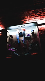 A store display featuring a variety of Lady Vans merchandise. Mannequin legs with colorful socks and shoes are prominently displayed. The background consists of other accessories and products arranged in an organized manner. The lighting is dark with neon and backlit illumination enhancing the visual appeal.