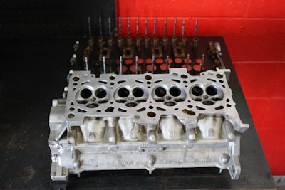 A metal engine cylinder head placed on a dark surface with a red wall in the background. The cylinder head features multiple circular openings and intricate metalwork, indicative of mechanical precision. In the background, various engine components like valves and springs are lined up neatly, suggesting a workshop environment.