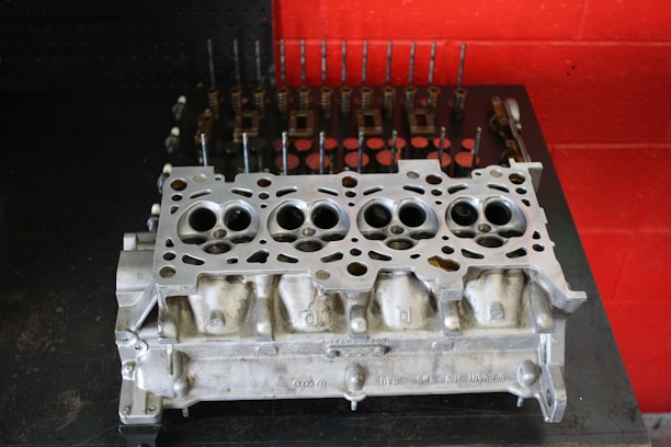 A metal engine cylinder head placed on a dark surface with a red wall in the background. The cylinder head features multiple circular openings and intricate metalwork, indicative of mechanical precision. In the background, various engine components like valves and springs are lined up neatly, suggesting a workshop environment.