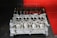 A metal engine cylinder head placed on a dark surface with a red wall in the background. The cylinder head features multiple circular openings and intricate metalwork, indicative of mechanical precision. In the background, various engine components like valves and springs are lined up neatly, suggesting a workshop environment.