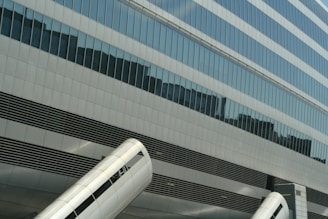 Glass building facade with reflective windows and large metallic cylindrical structures in the foreground. The geometric pattern of the windows and the metallic surfaces create an urban, modern feel.