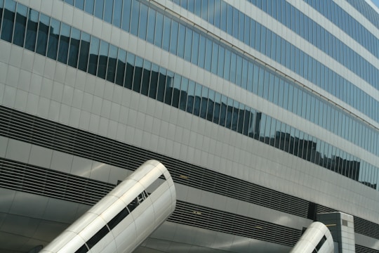 Glass building facade with reflective windows and large metallic cylindrical structures in the foreground. The geometric pattern of the windows and the metallic surfaces create an urban, modern feel.