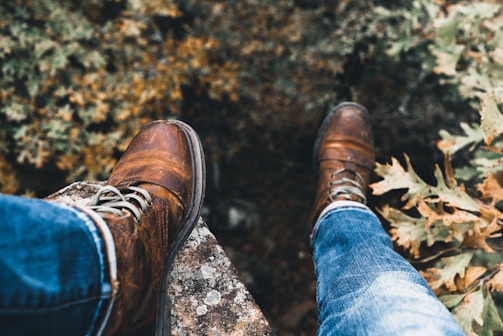 A pair of brown leather boots resting on a weathered rock edge. The boots are worn with denim jeans. Surrounding foliage has autumn colors, with hints of yellow and brown leaves.