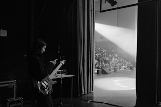 A musician holding a guitar stands backstage, looking at a phone. In the background, a crowd is visible in a partially lit auditorium. Stage lights shine toward the seating area, creating a stark contrast between the dimly lit backstage and the bright stage.