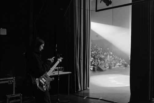 A musician holding a guitar stands backstage, looking at a phone. In the background, a crowd is visible in a partially lit auditorium. Stage lights shine toward the seating area, creating a stark contrast between the dimly lit backstage and the bright stage.
