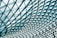 An intricate geometric pattern composed of triangular glass panels forms a modern architectural ceiling. The panels are supported by a metal framework, creating a dynamic grid that captures light beautifully.