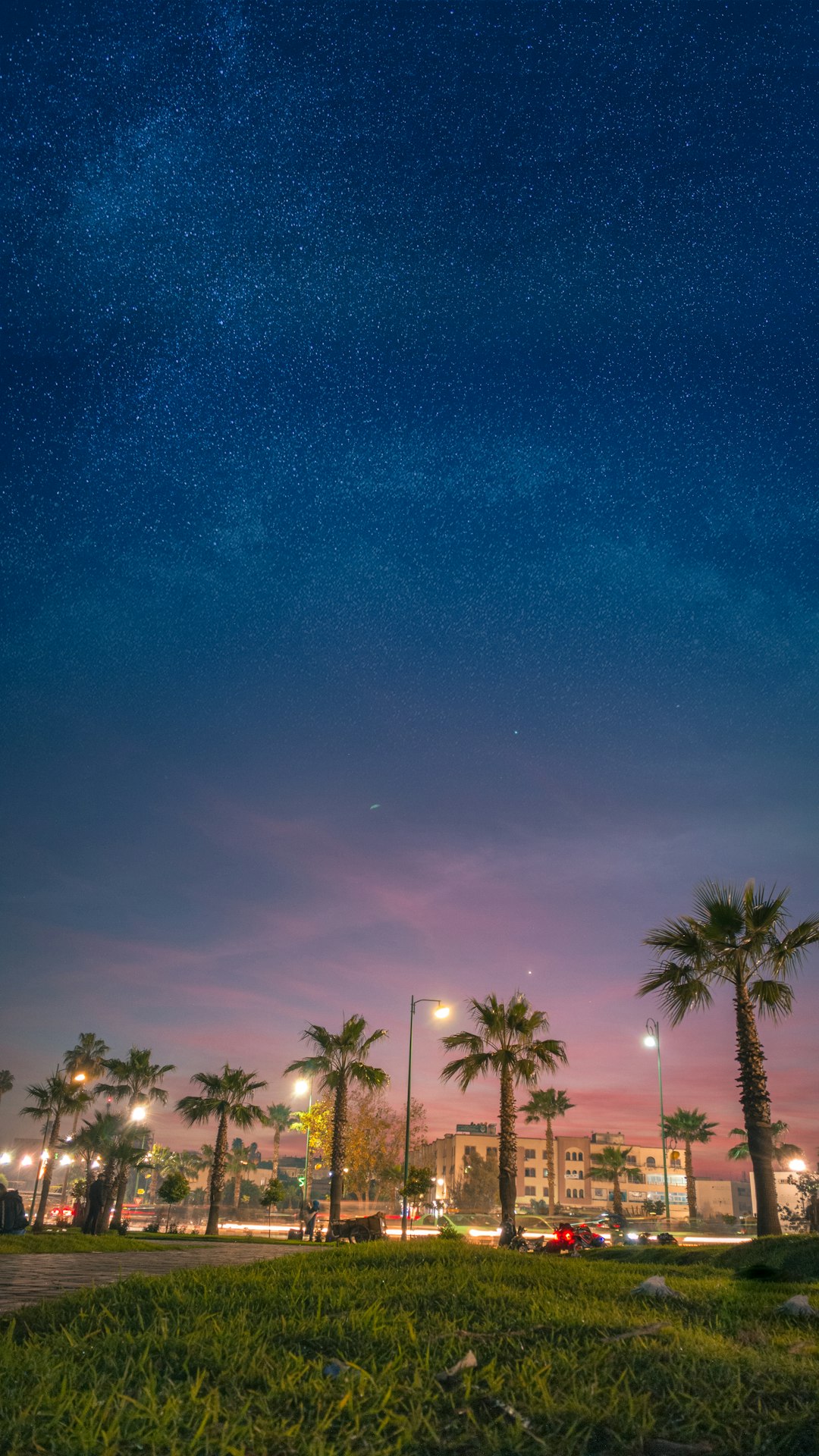 vehicles on green field near trees viewing buildings and hotels under blue and orange sky during night time