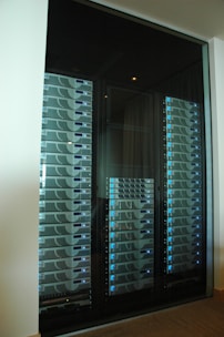 A server room or data center featuring several racks of network servers with blue indicator lights. The setup is organized and enclosed within a glass cabinet.